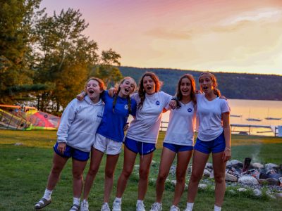 Group of girls bonding by a campfire at dusk, illustrating the lifelong friendships formed during a 4-week Maine summer session.