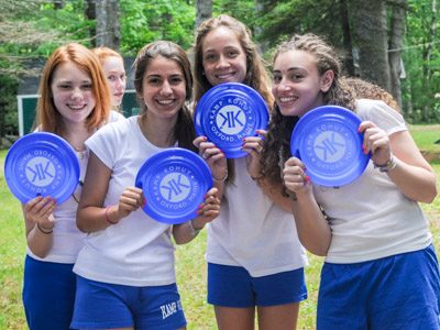 A group of smiling girls wearing matching blue shirts and white shorts, holding frisbees featuring the Kamp Kohut logo.